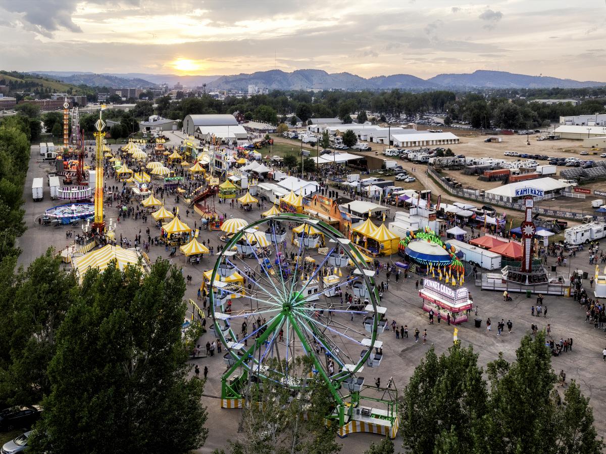 Aerial view of a bustling fairground at sunset, featuring a large Ferris wheel, colorful tents, and various rides. People are scattered throughout the vibrant scene, adding a lively atmosphere.