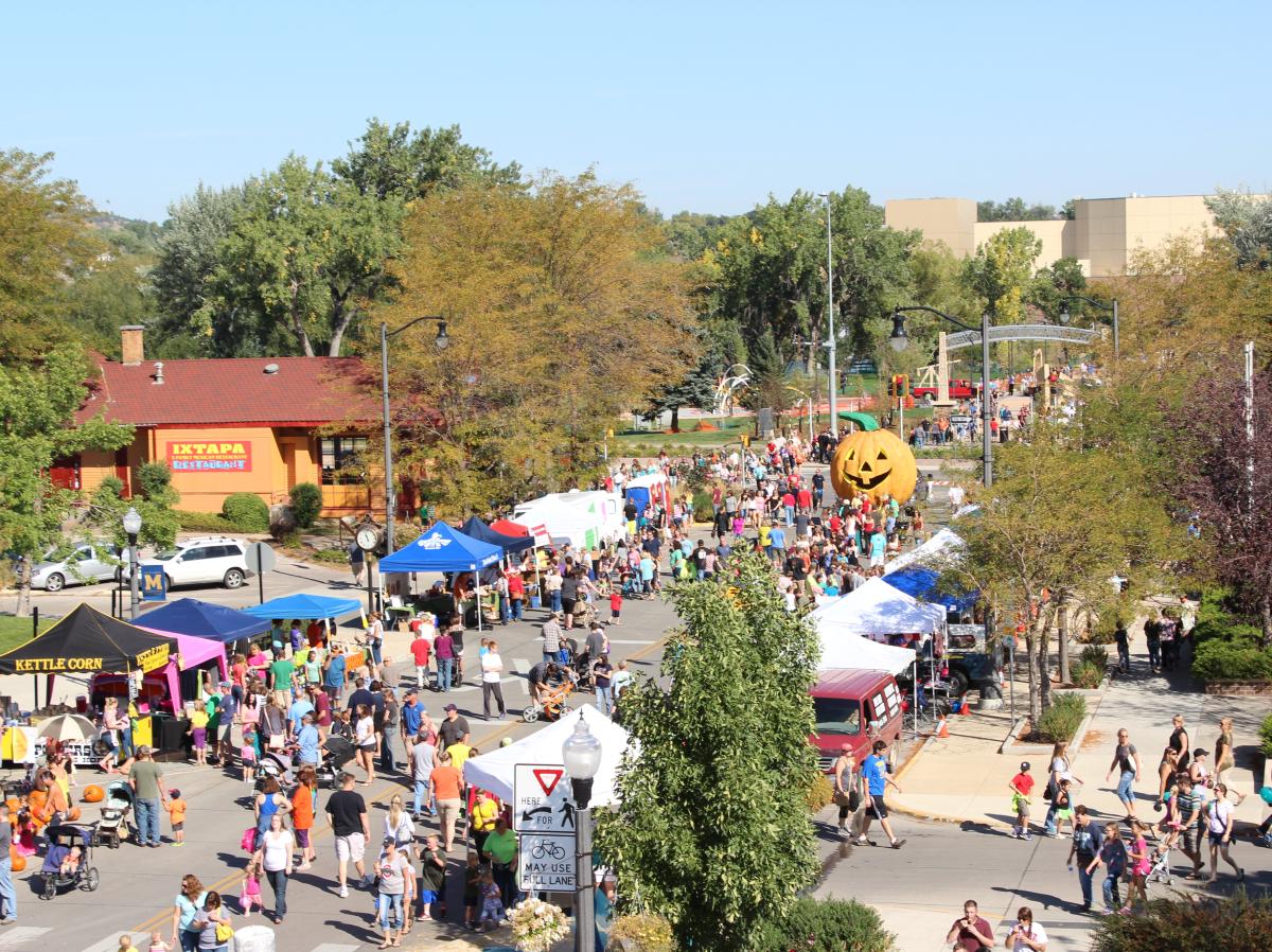 A bustling fall festival with colorful vendor tents, crowds of people, and a large inflatable pumpkin. Trees with autumn foliage and a sunny sky convey a festive, lively atmosphere.