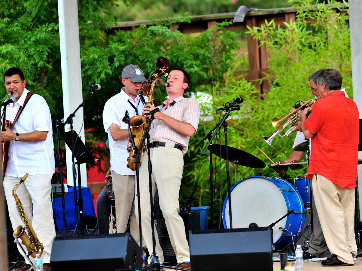 Musicians perform an outdoor concert at Columbia Street Landing in Covington, with saxophones, trumpet and drums onstage.