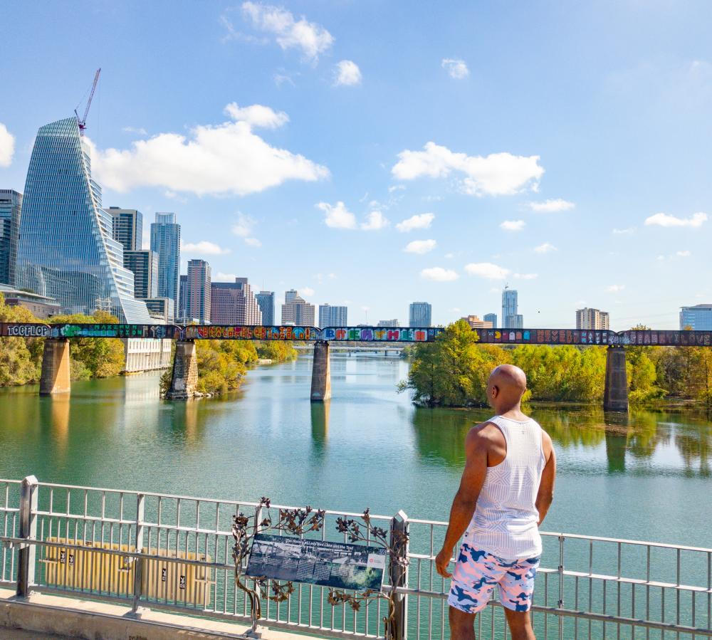 Man walking on Pfluger Pedestrian Bridge on the Hike & Bike Trail, looking across the water at the downtown skyline.