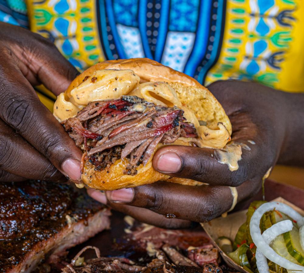 Photo of man's hands holding a brisket sandwich towards the camera at La Barbecue