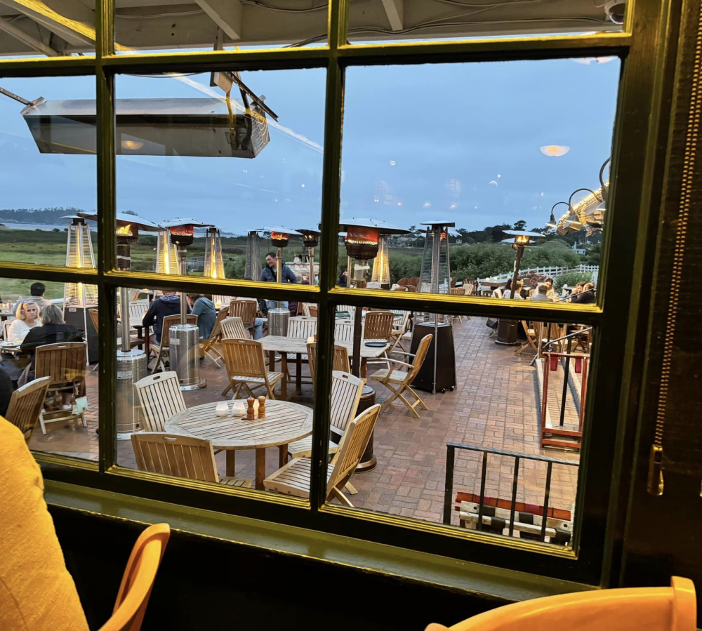 This is an image of the outdoor patio tables at the restaurant at Carmel Mission Ranch in Carmel Valley taken from an indoor table. The wooden tables and chairs are surrounded by heat lamps, creating a warm ambiance. The ocean can be seen from a distance.