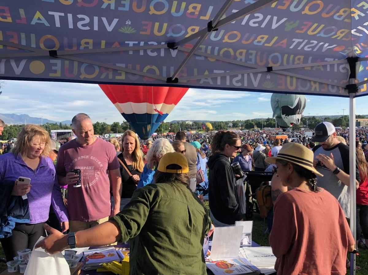 Two Visit Albuquerque employees work a booth at Labor Day Liftoff with a crowd of people in the background
