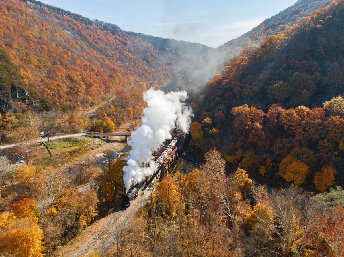 A steam train travels through a mountain valley surrounded by vibrant autumn foliage.