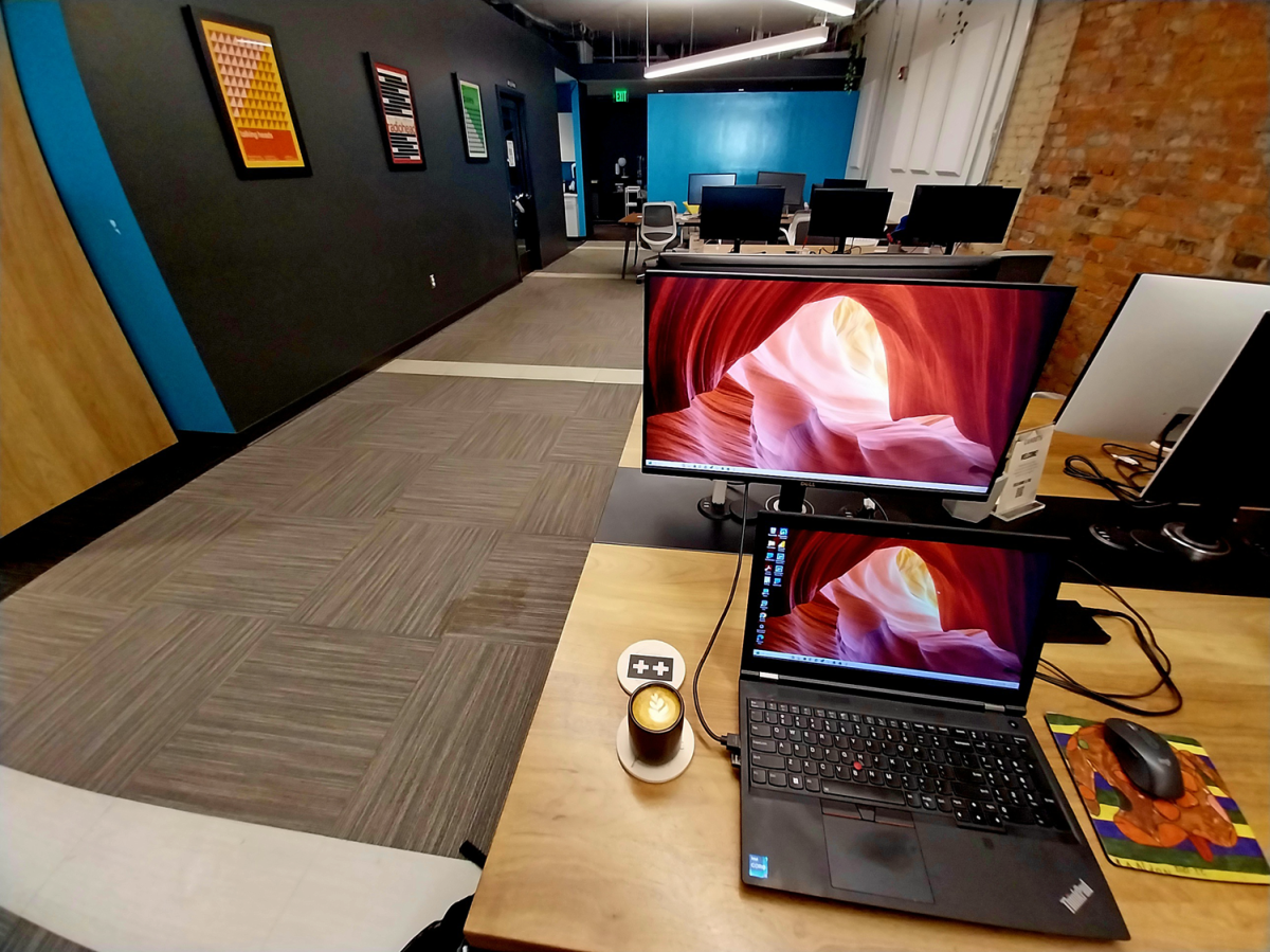 laptop and coffee sit on table hooked up to another monitor. Past the table is a long hallway leading to more rooms and computer tables.