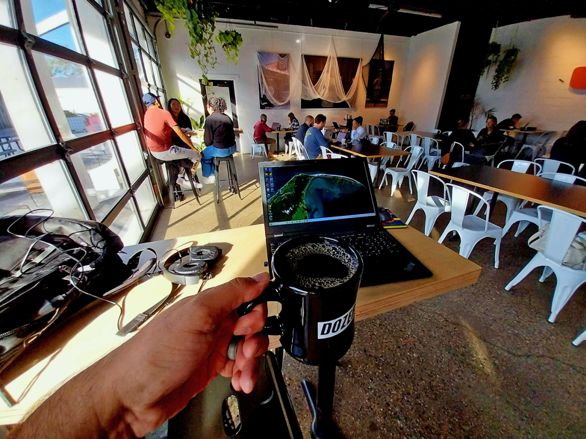 Person holds coffee mug in front of themselves, with a laptop sitting on a table behind the cofee mug. People are chatting, sitting at tables in the background.