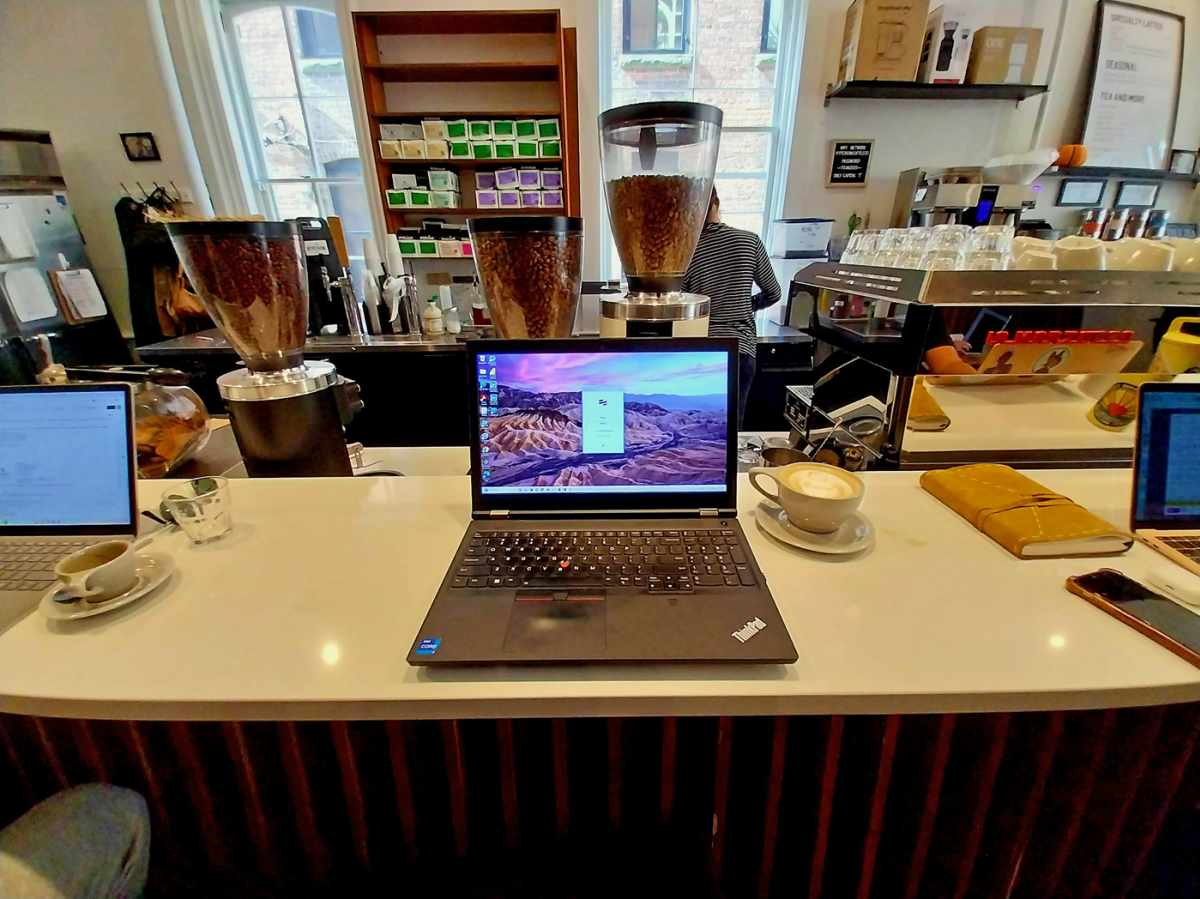 A laptop sits in the center of a counter with a barista making a coffee drink in the back.