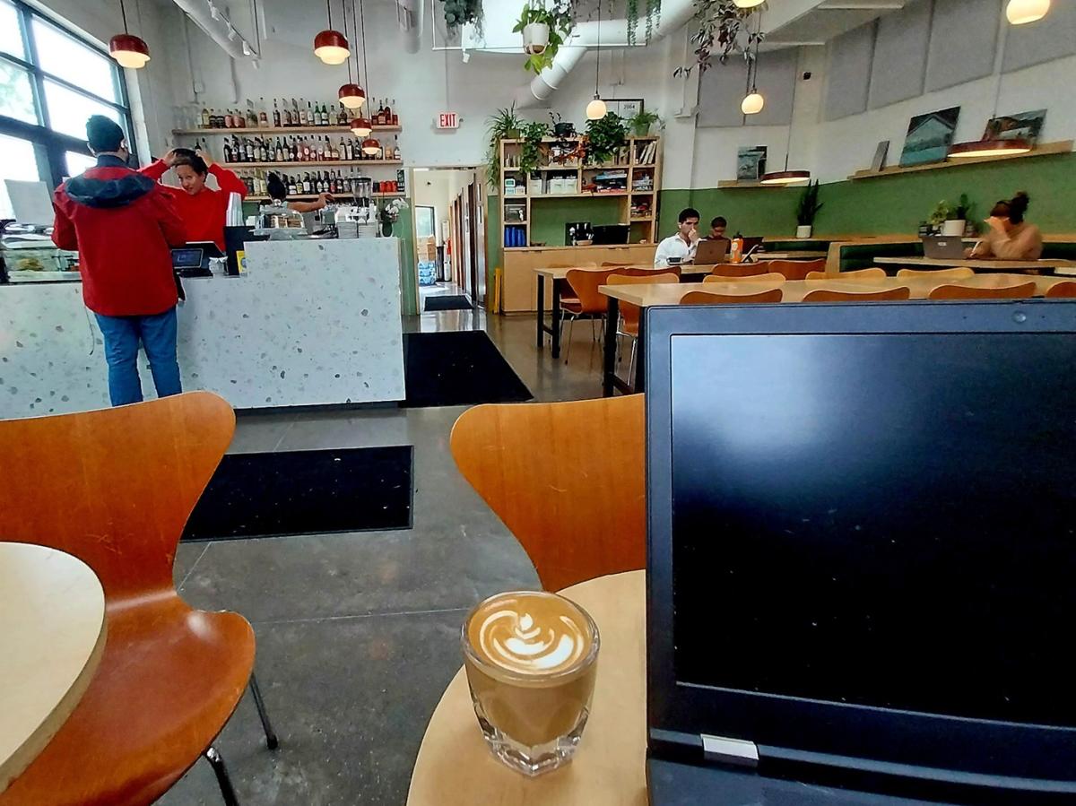 Laptop and coffee sit on a table as someone in the distance orders a beverage at the cafe bar.