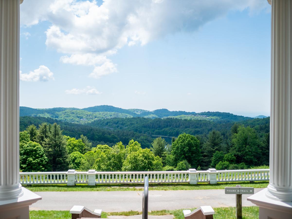 A beautiful view of a blue sky and tree-covered rolling Appalachian mountains can be seen between two white colonial style columns at Moses Cone Manor.