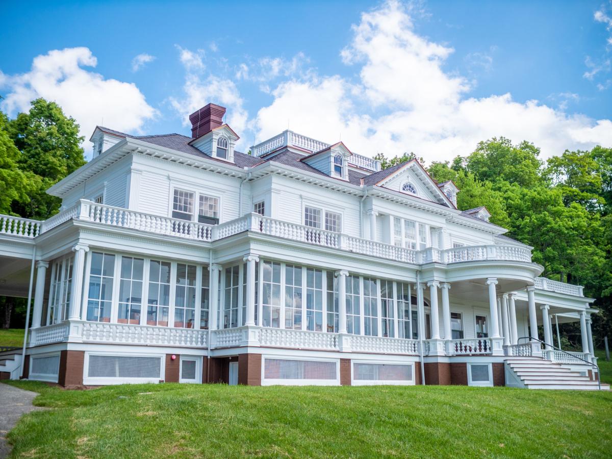 A large white manor with columns and a wrap-around porch on the first and second levels.