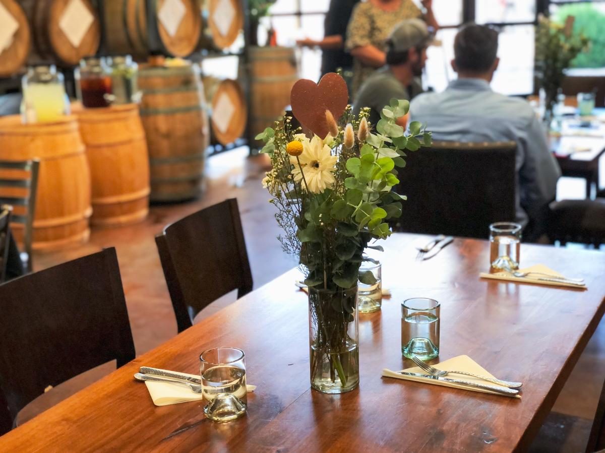 Large wood rectangle table with black metal chairs around it. Glasses with water, silverware, and white napkins are at each place setting. The centerpiece is a tall glass vase with green and white florals inside. In the background distilling barrels can be seen as well as two men sitting another identical table.