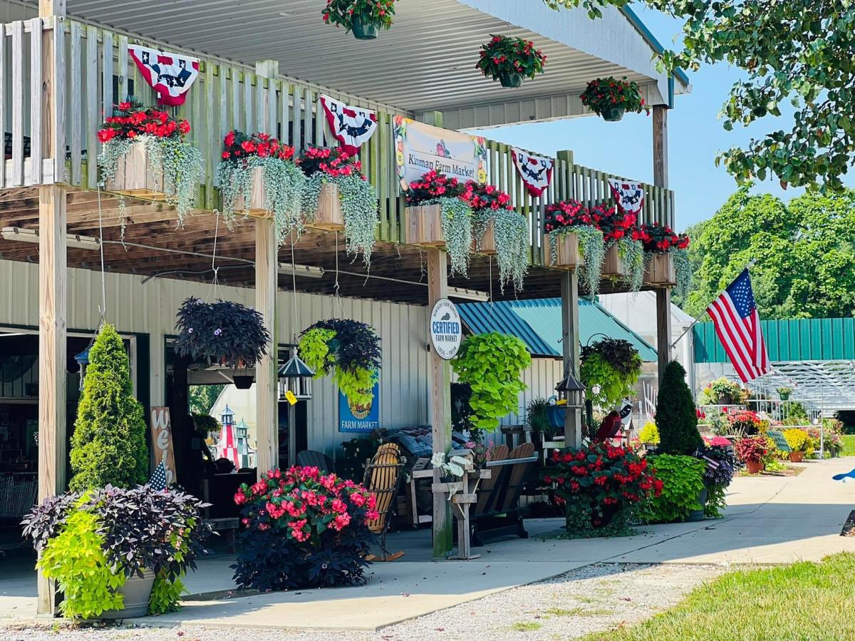 Image is of a building on the Kinman Farm Market grounds with flowers hanging on the balcony and on the floor with a sign that say's "Kinman Farm Market".