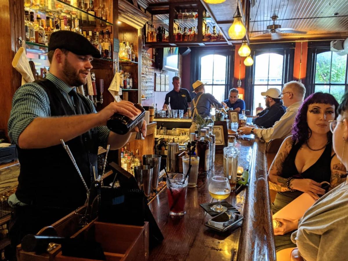 Men and women sitting at a wood bar while a bartender in a cap pours a cocktail.