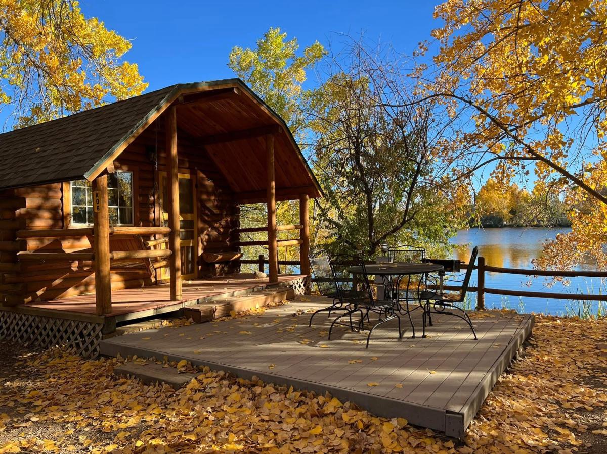 A cabin with a lake in the background. The trees around the cabin have yellow leaves and there are leaves on the ground