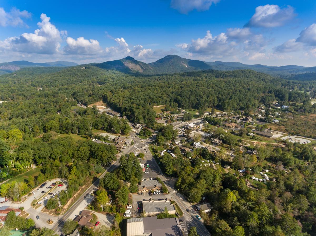 A high-angle aerial view of the small town of Cashiers, North Carolina. The town's central intersection and buildings are nestled within a dense green forest, with two prominent mountain peaks rising in the background under a blue sky with scattered clouds.