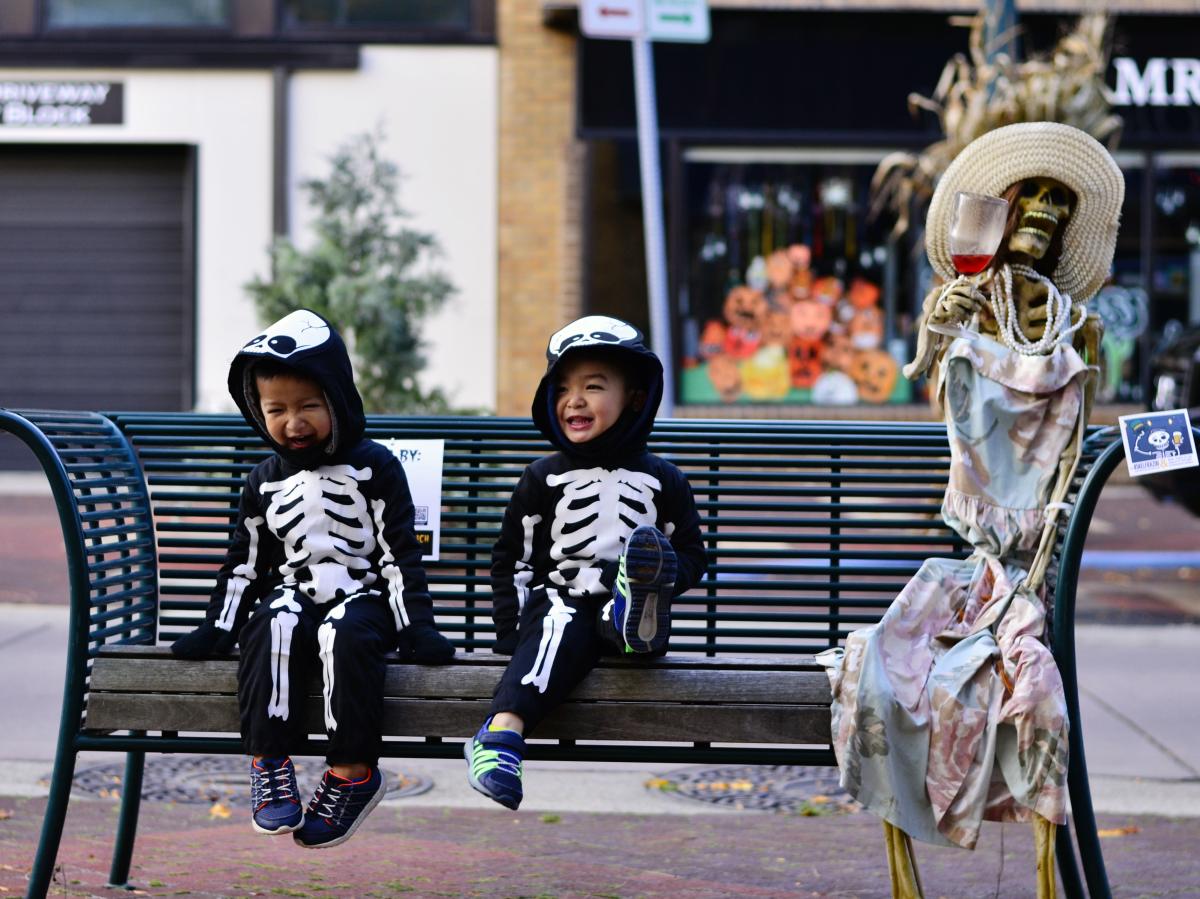 2 kids in skeleton costumes sitting on a bench next to a dressed up skeleton