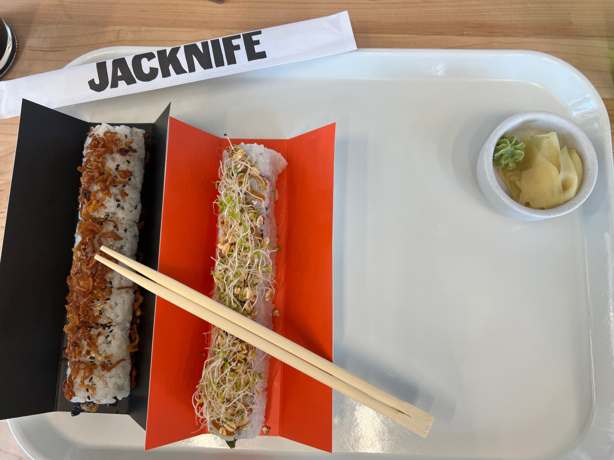 Two rolls of sushi, one topped with crispy shallots and the other topped with sprouts, sit on a tray next to chop sticks and a small bowl of wasabi and ginger at Jacknife