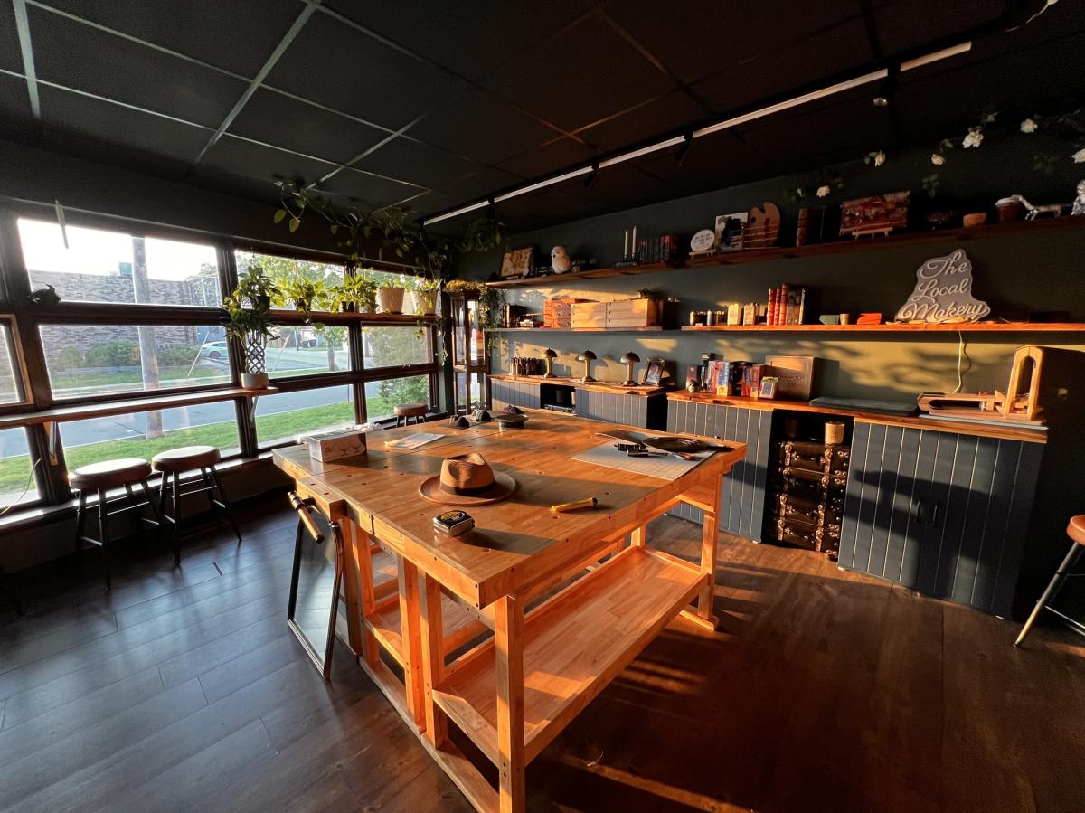 A warmly lit workshop-style interior with a large wooden worktable in the center, shelves of tools and materials along the wall, dark wood floors, and a wide window letting in natural light. The space feels organized, rustic, and creative.