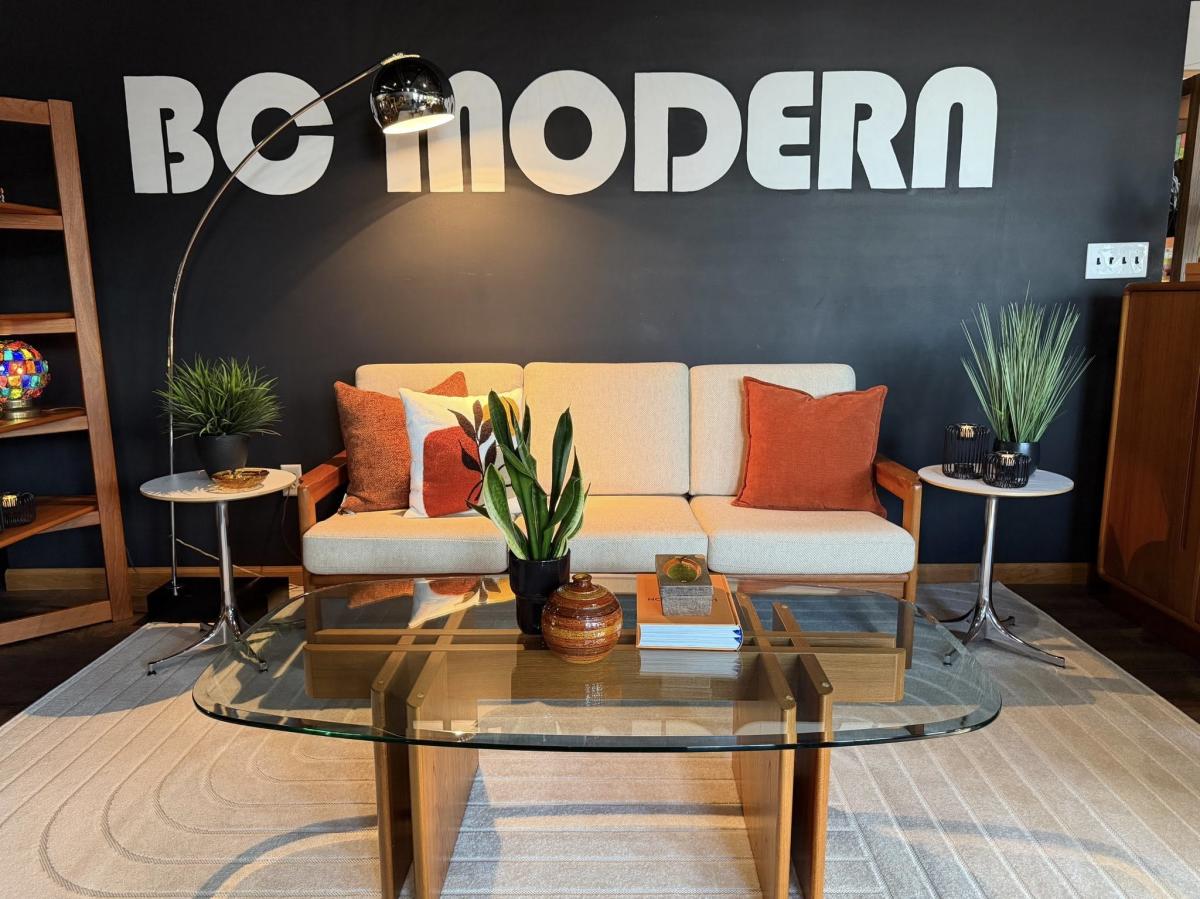 Mid-century modern showroom with a cream sofa and rust-colored pillows set against a dark wall reading “BC Modern,” featuring a glass coffee table, potted plants, side tables, and a curved floor lamp.