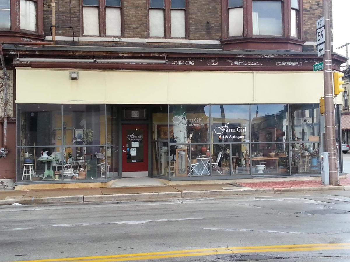 The storefront of Farm Girl Art & Antiques occupies a historic corner building with large display windows showcasing vintage furniture, home décor and antiques. A red entrance door sits between the window displays along a city street.
