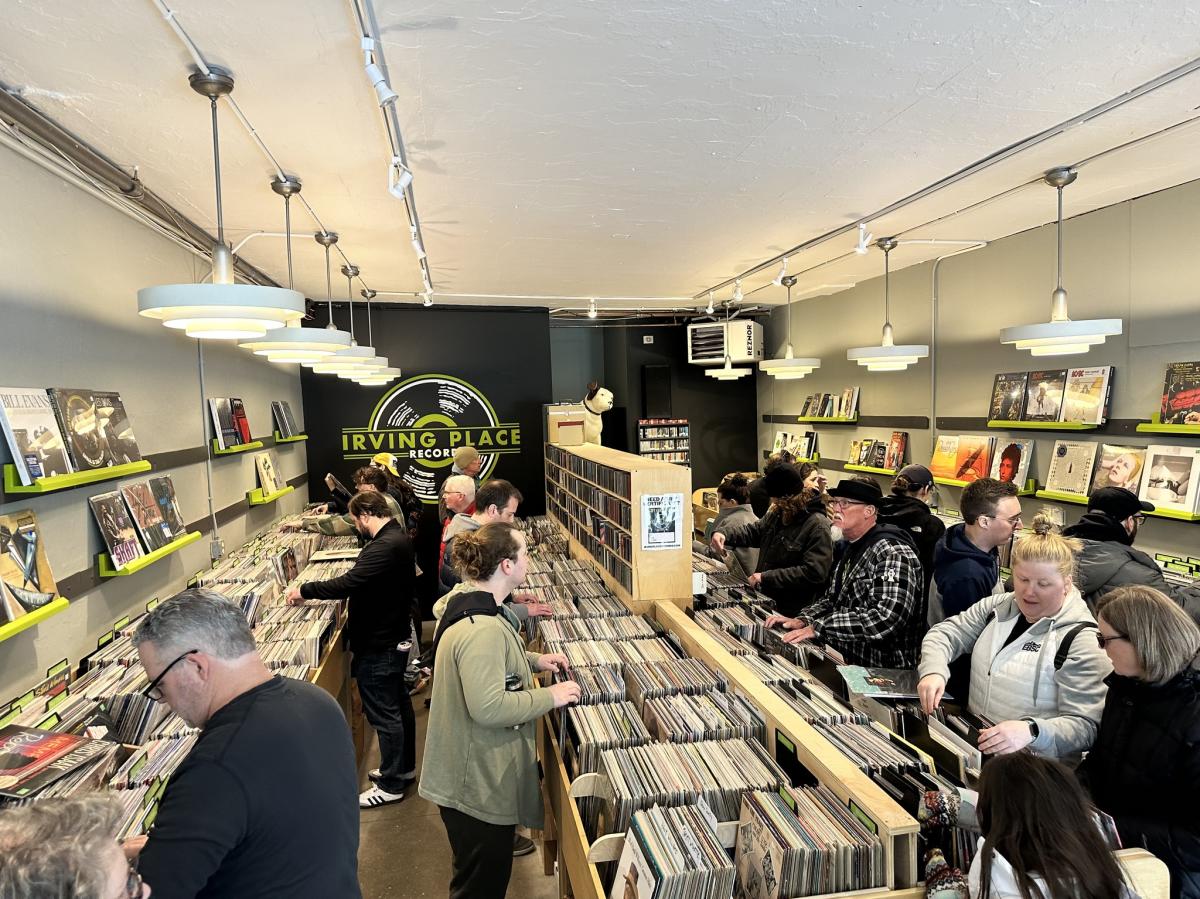 A busy record store is filled with people browsing tightly packed rows of vinyl, flipping through albums and comparing finds. Bright overhead lights and clean, modern shelving line the space, with a large “Irving Place Records” sign on the back wall anchoring the scene.