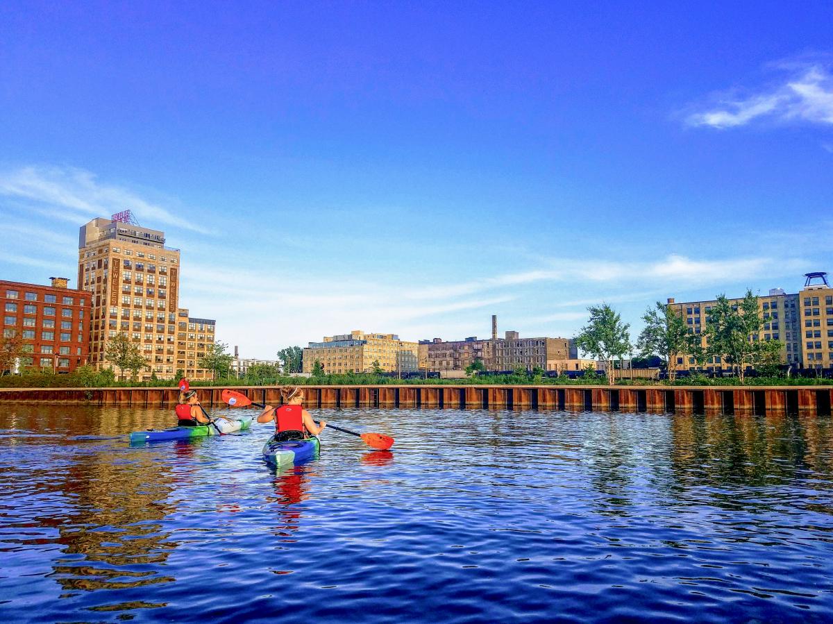 Two kayakers on the Menomonee River with calm waters and and urban backdrop