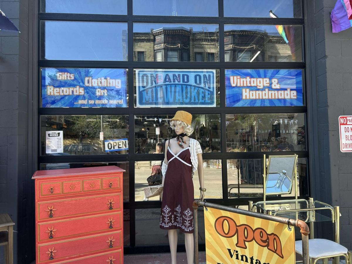 The entrance to On and On Milwaukee features large glass garage-style doors with colorful signs advertising vintage, handmade goods, records, clothing, art and gifts. A mannequin in vintage clothing, a bright red dresser and other furniture are displayed outside the shop beneath Pride flags.