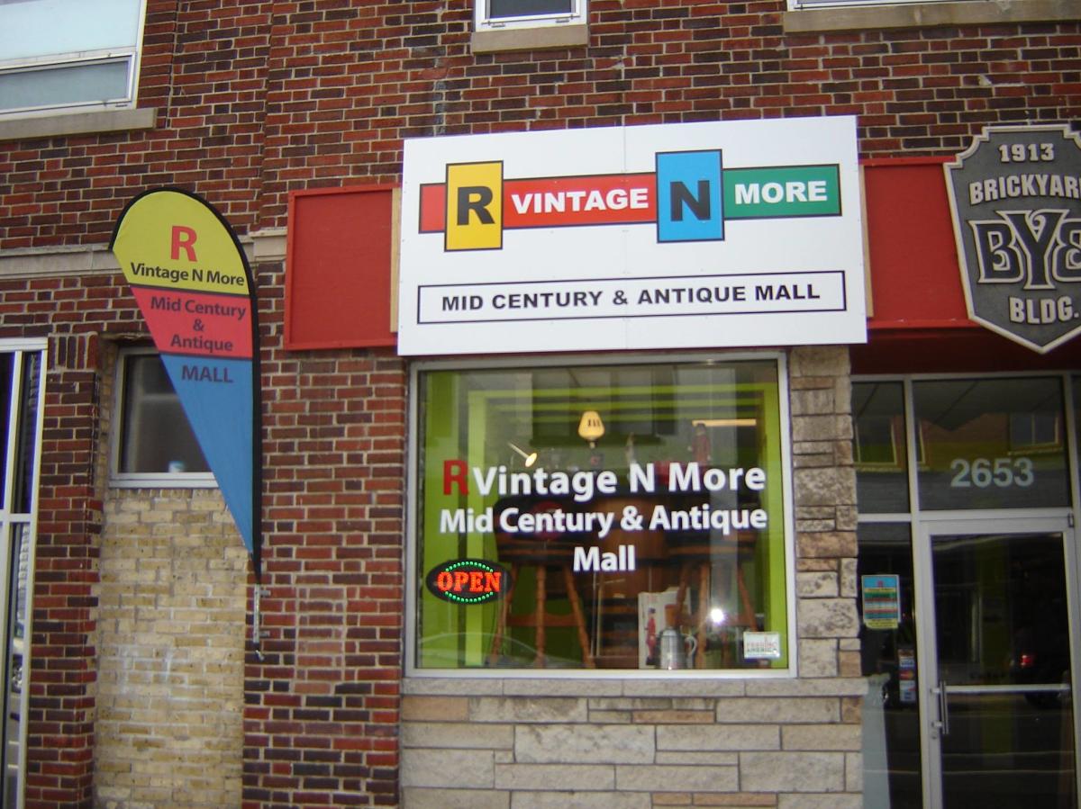 Brick storefront for R Vintage N More Mid Century & Antique Mall with colorful signage, large front window display, and an illuminated “Open” sign.