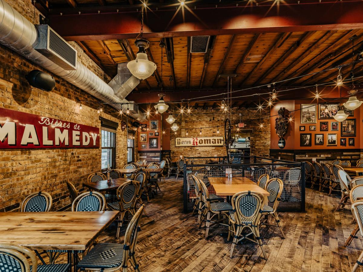A warm, rustic restaurant interior with exposed brick walls, wooden beams, vintage signage, and neatly arranged tables and chairs.