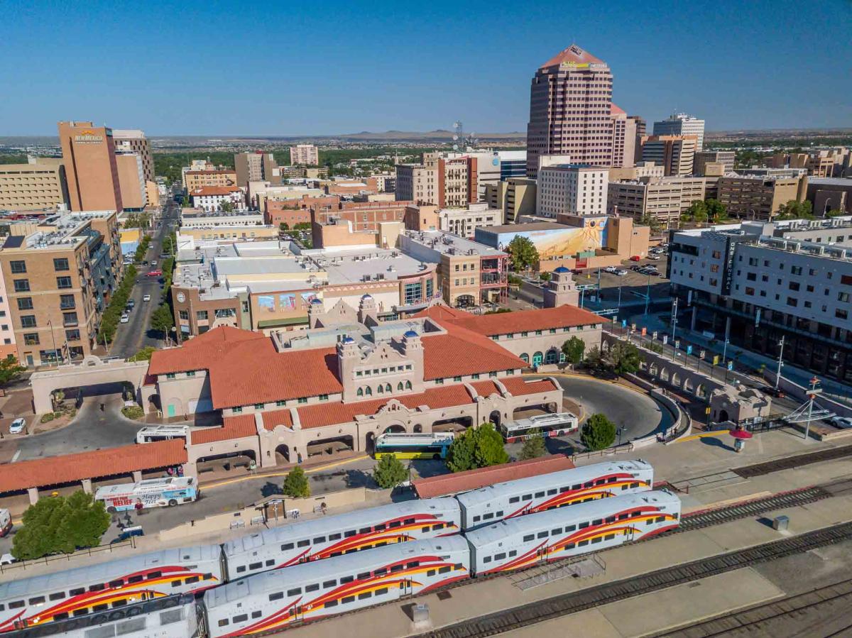 Aerial view of Albuquerque's Alvarado Transportation Center with multiple New Mexico Rail Runner trains at the platform, surrounded by city buildings under a clear sky.