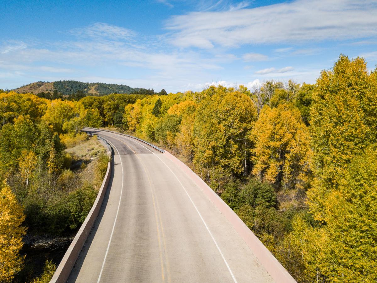 A scenic road curves through a landscape filled with trees displaying vibrant autumn colors. The sky is mostly clear with some clouds. Rolling hills are visible in the background.