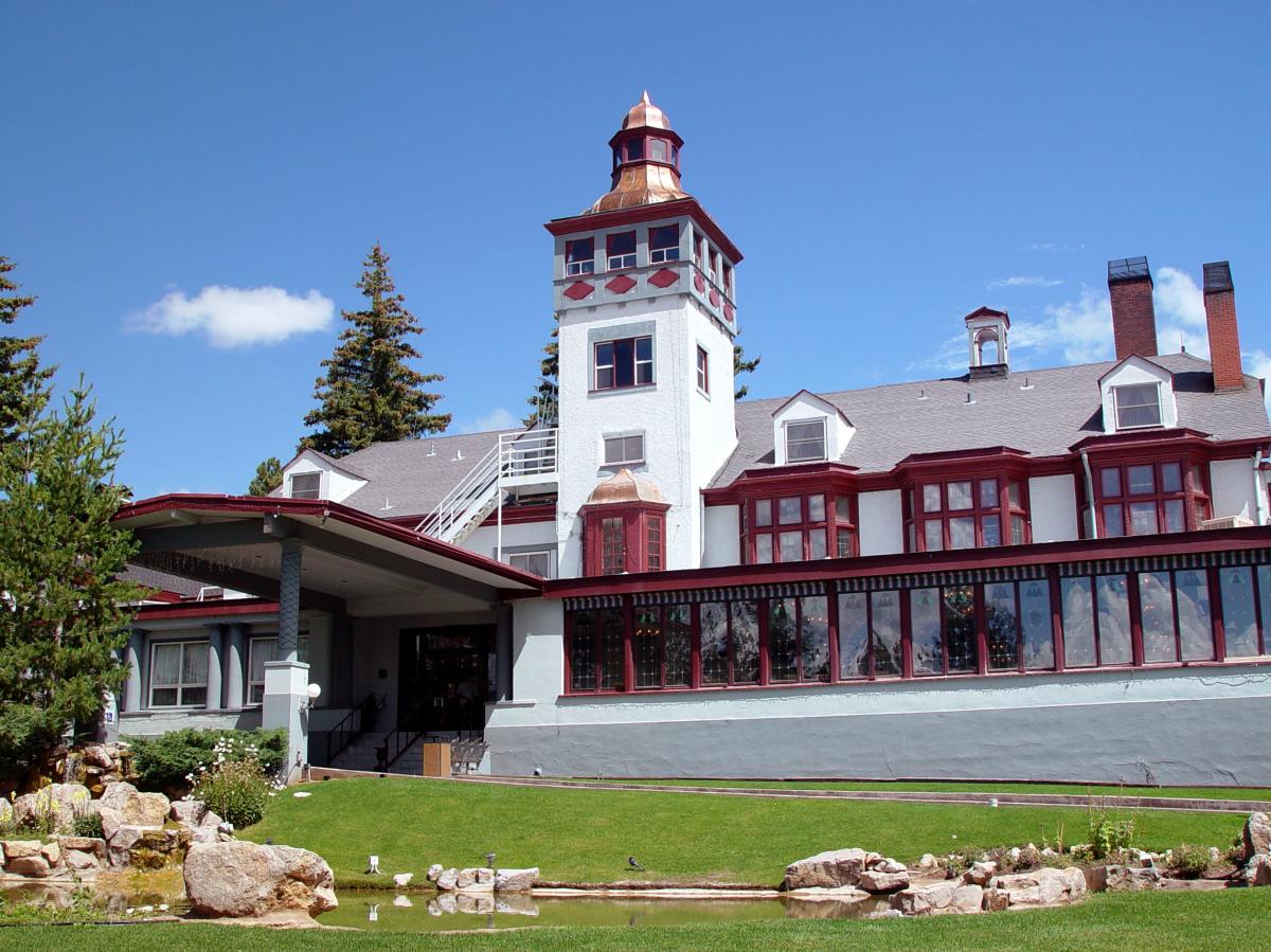 View of the The Lodge at Cloudcroft on a sunny day, featuring a prominent red and white building with a distinctive tower and surrounded by lush greenery and a small pond in the foreground.