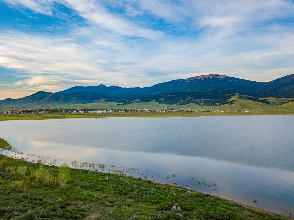 Aerial view of a serene lake with a small town nestled at the base of expansive mountains under a partly cloudy sky.
