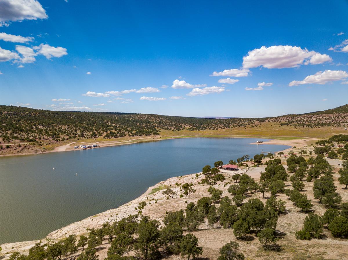 Aerial view of Bluewater Lake State Park with clear blue waters, surrounded by rugged terrain and sparse vegetation under a bright sunny sky.