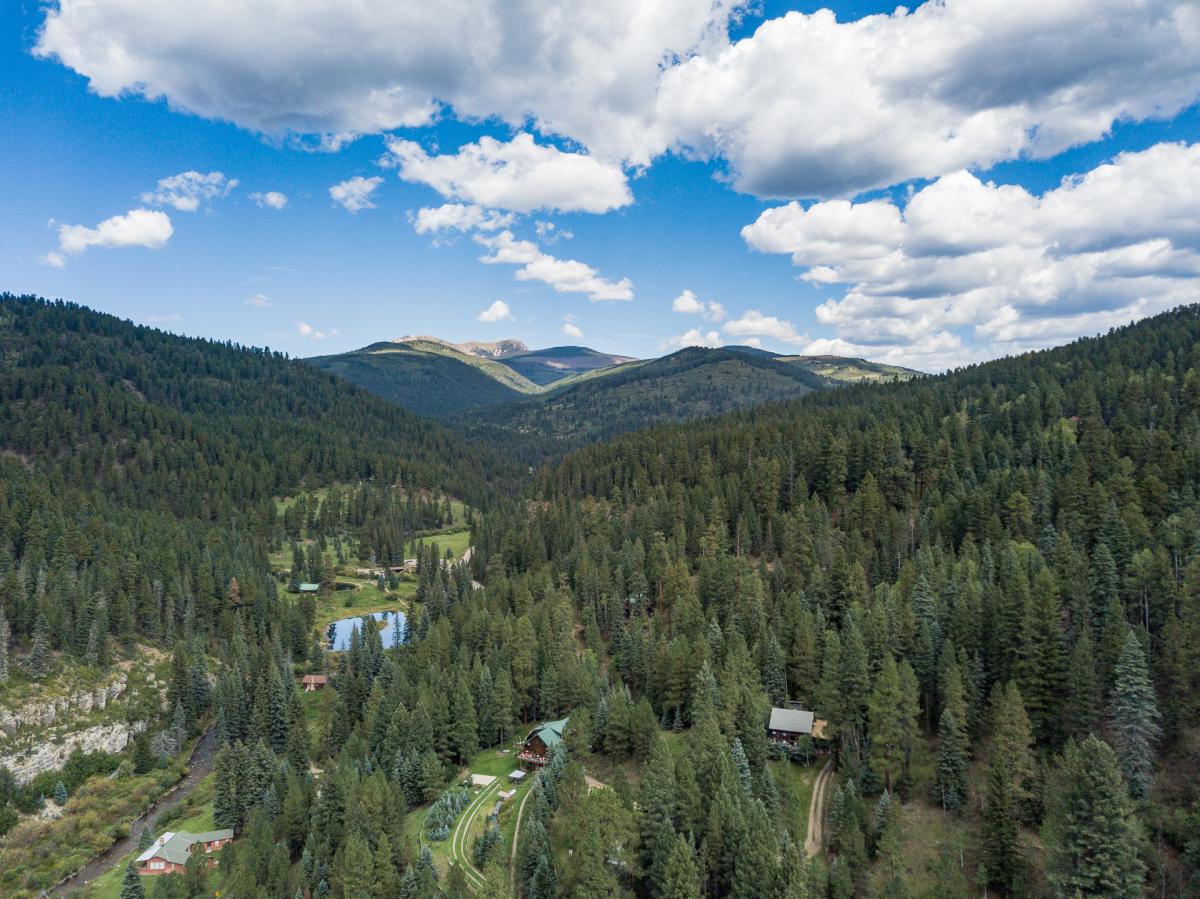 Aerial view of a lush, forested mountain landscape with a small body of water and several cabins and structures nestled among the trees. The sky is partly cloudy, and distant mountains are visible on the horizon.