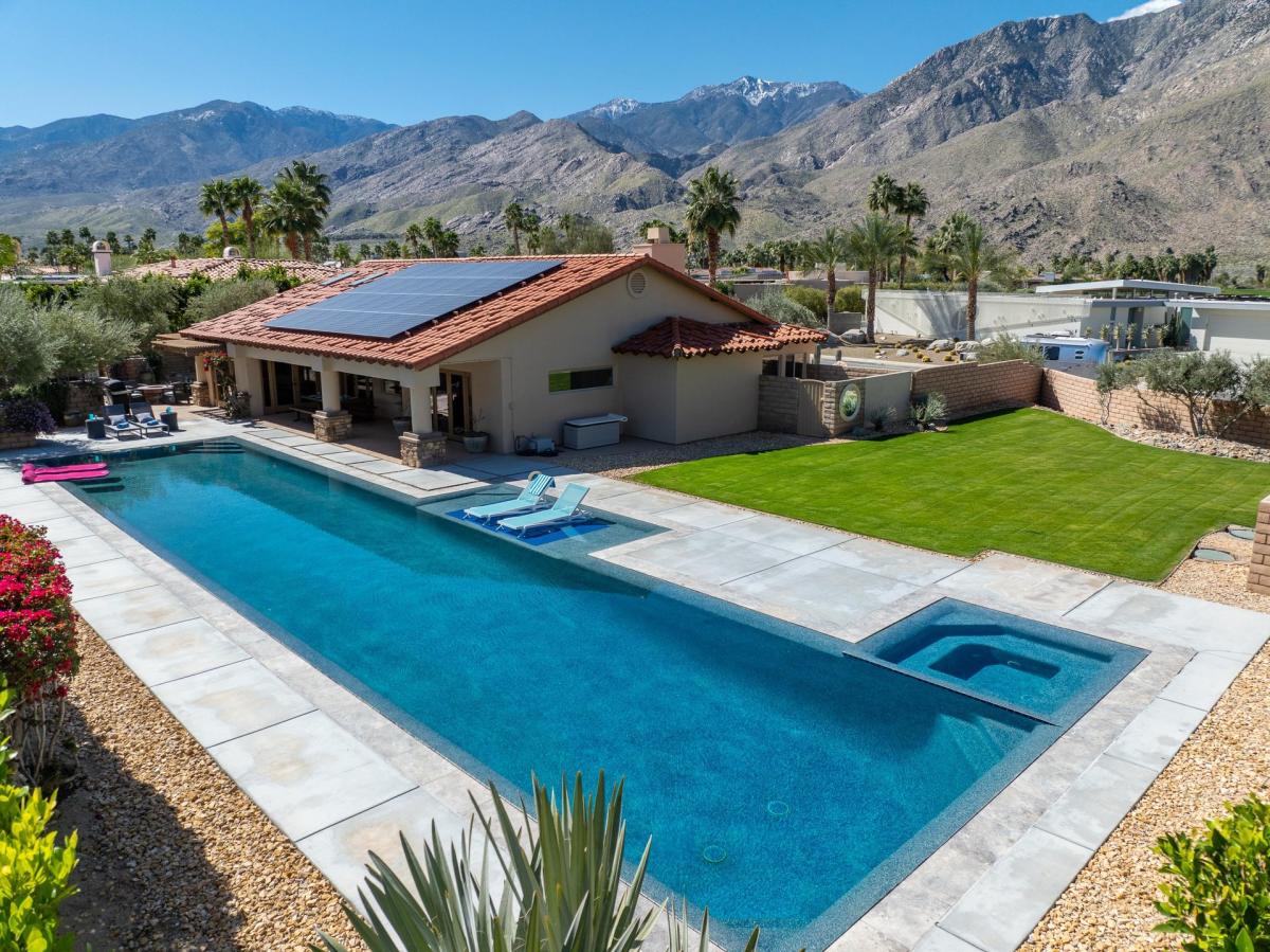 Aerial view of a modern single-story house with a red tile roof, equipped with solar panels, next to a large swimming pool, with mountains in the background.