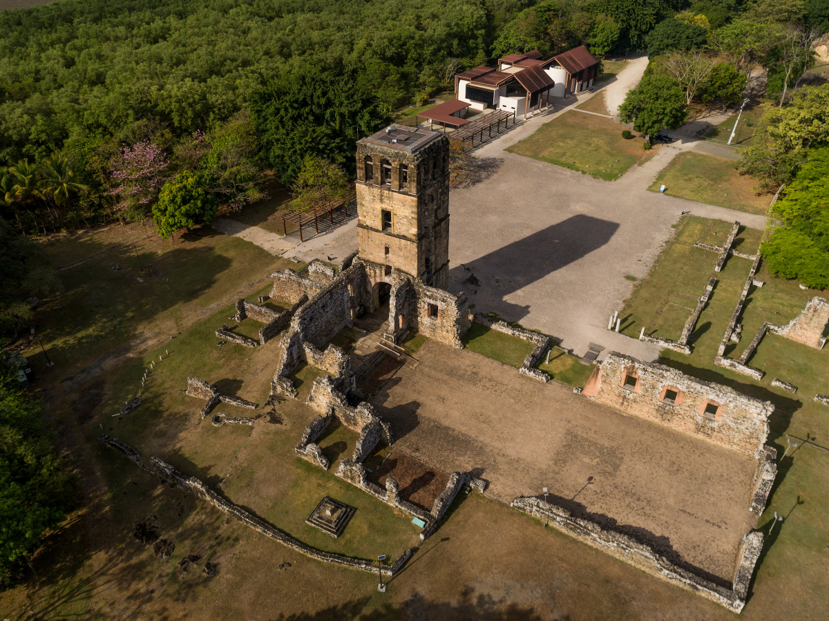 The image shows an aerial view of an ancient archaeological site. The site appears to be a large complex of stone ruins, with a prominent bell tower standing tall in the center. The ruins are surrounded by lush green vegetation, suggesting a tropical or subtropical climate of Panama City