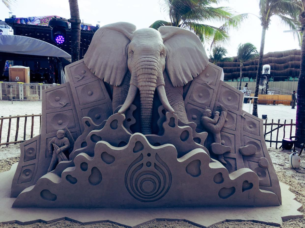 Sand sculpture showing an elephant coming towards the camera. Carved on either side of the elephant are speakers and small people, and a shape in front.