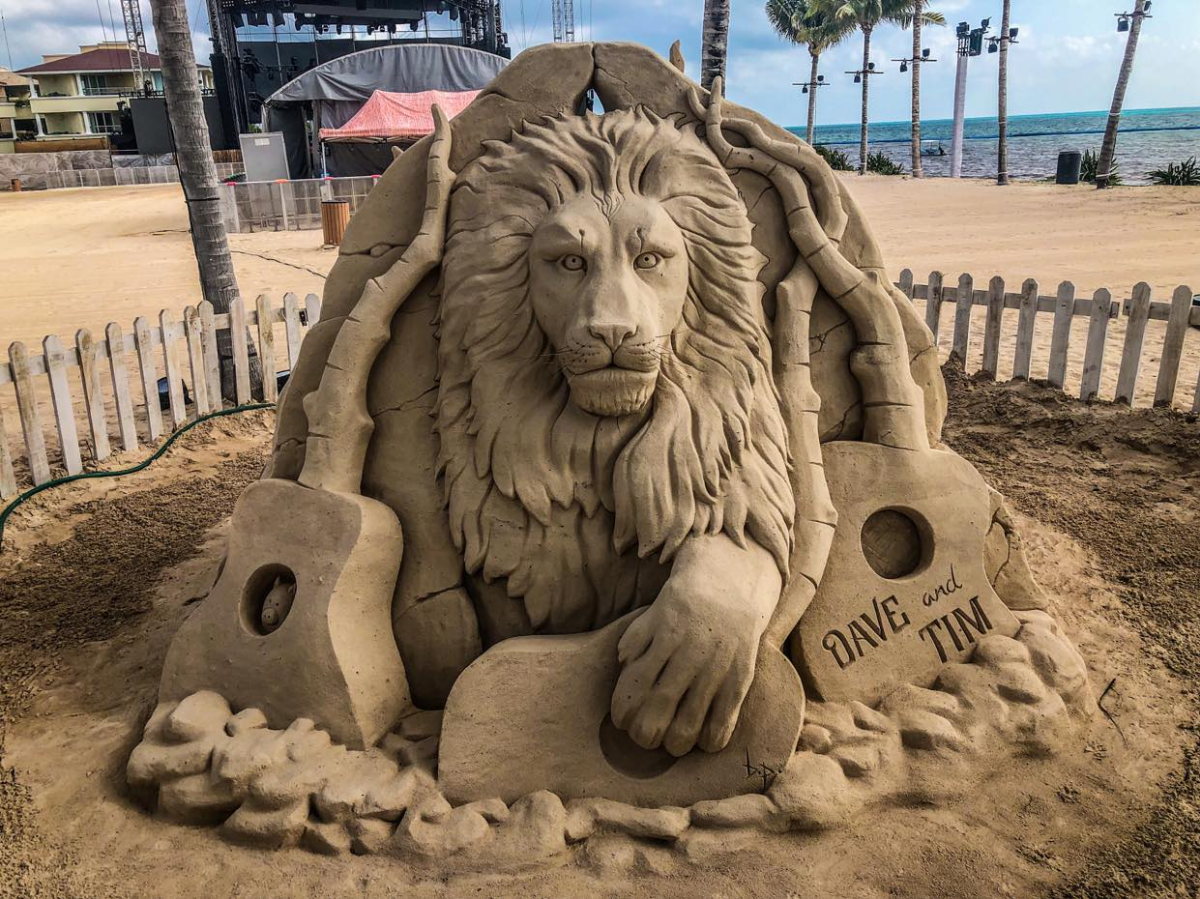 Sand sculptor surrounded by a fence that shows a lion surrounded by guitars that seem to be made of bone or part of hte ground.