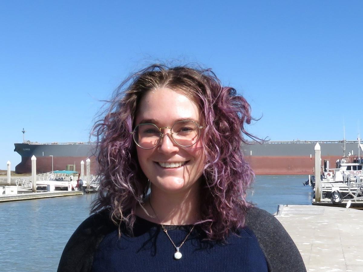 Woman with purple curly hair stands in front of a ship channel and smiles at the camera