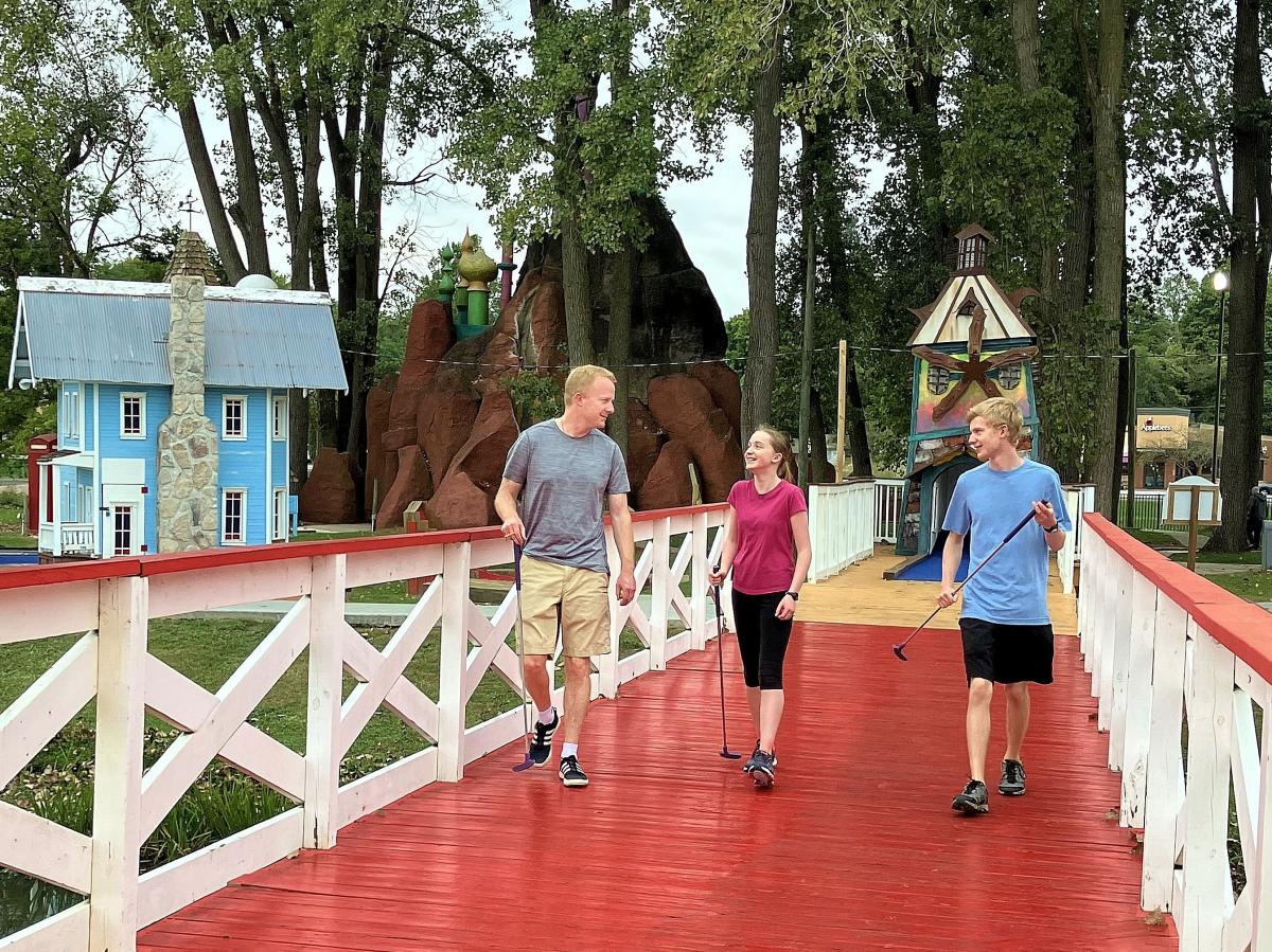 A father and his two teenage children walk along a bridge while holding golf clubs.