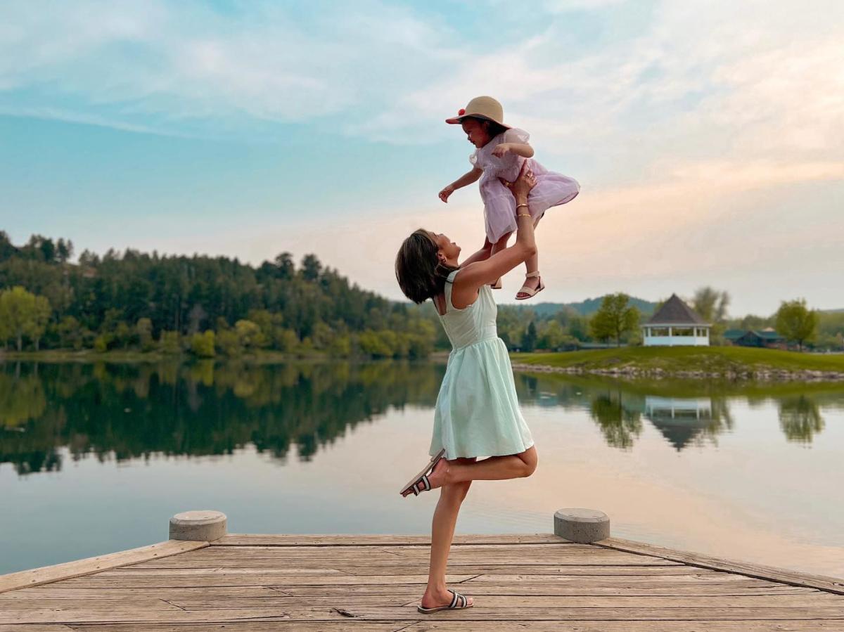 A woman in a light dress joyfully lifts a toddler in a pink outfit and sunhat on a lakeside dock, with serene water and a gazebo in the background at Canyon Lake Park