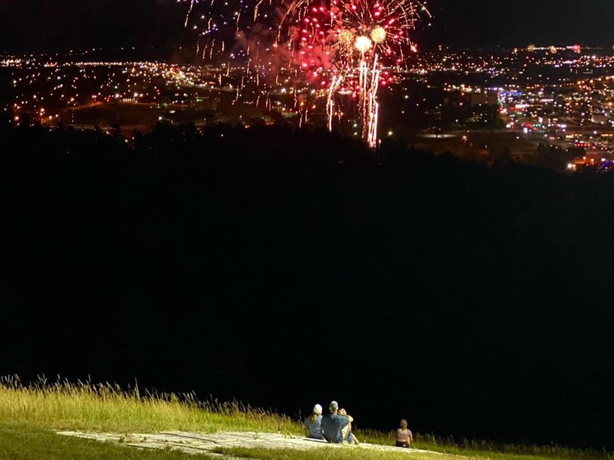 A family, illuminated by the lights on M Hill in Downtown Rapid City, watches the Rapid City city fireworks show with city lights sparkling on the horizon.