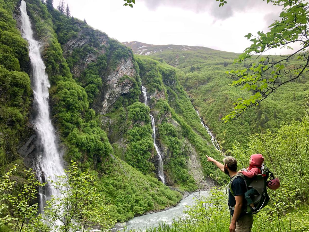 a man with a baby on his back points to three waterfalls in a lush canyon