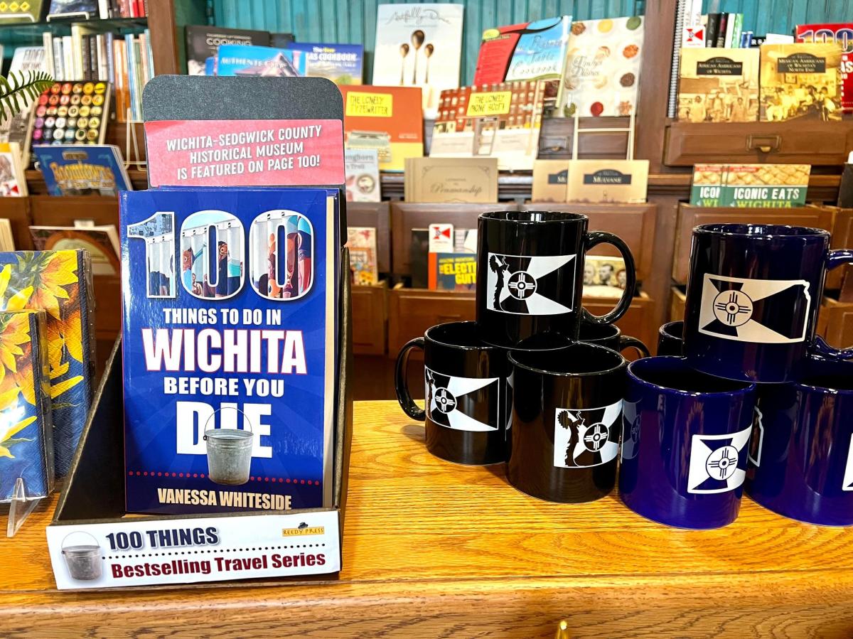 A photo of the book 100 Things To Do in Wichita Before You Die" next to coffee mugs with Wichita flag images at Watermark Books & Cafe in east Wichita