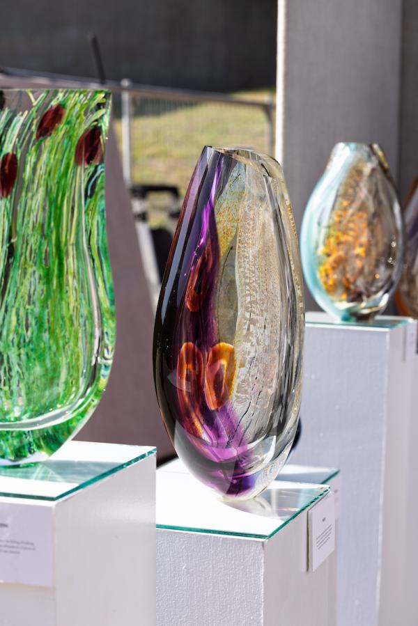Three colorful glass vases rest on pedestals at The Woodlands Waterway Arts Festival. The one closest to the viewer looks like it contains bright green strands of sea grass. The focal piece is bright white and purple with bright orange rings within in. The most distant piece is blue with flecks of orange and brown.