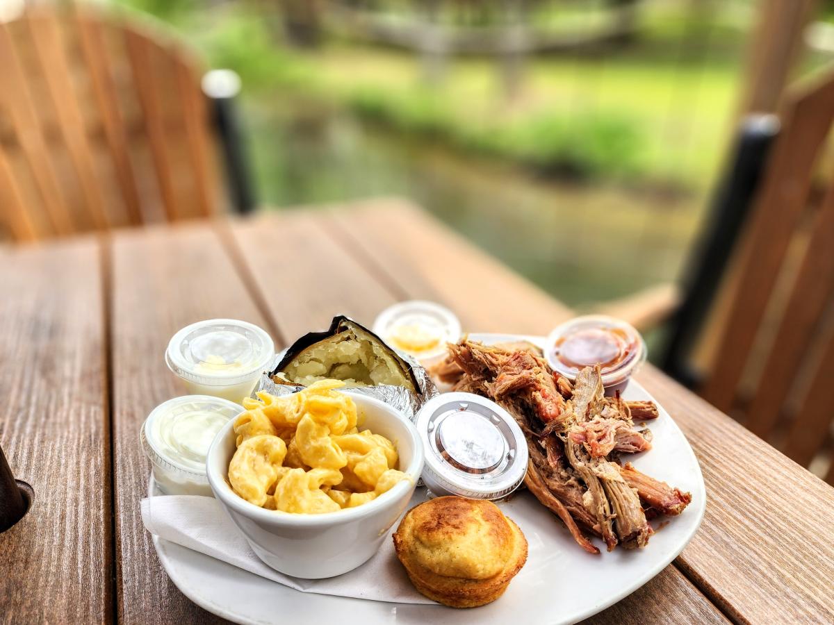 A plate of barbecue, baked potato, mac and cheese, and cornbread on a wooden picnic table.