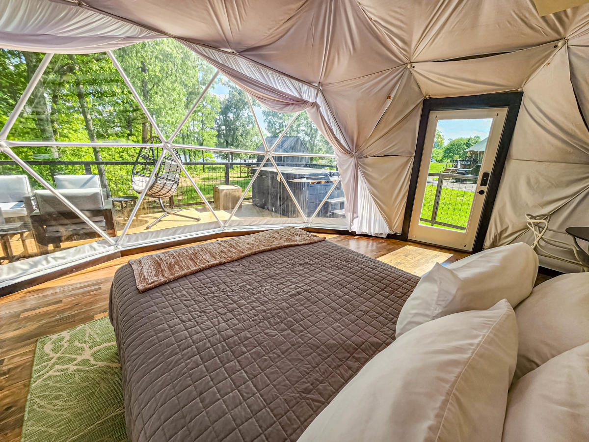 Inside the yurt at Starlight Haven on Weiss Lake. A clear window shows outside from a comfortable beige colored bed with pillows.