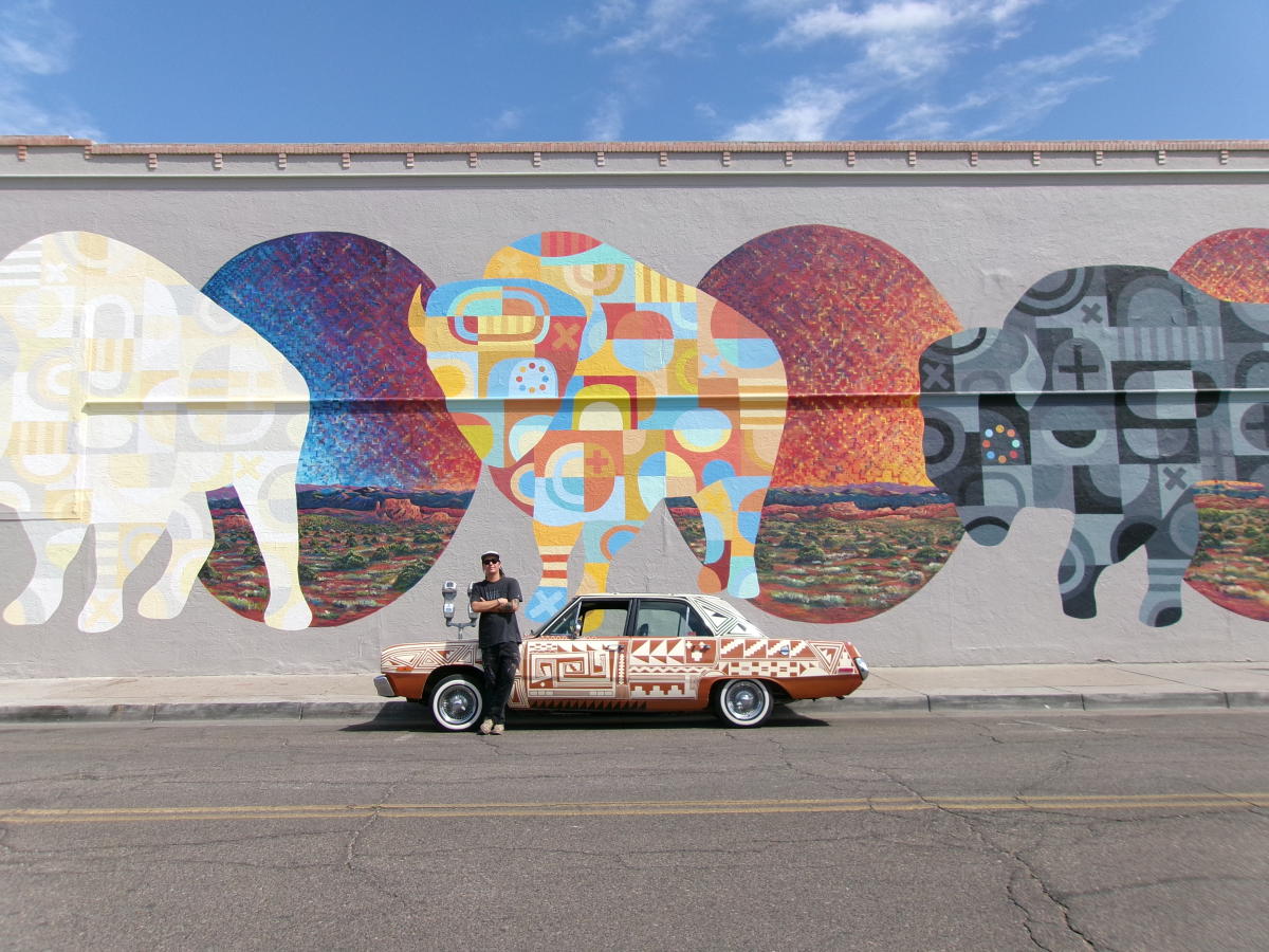 Jesse Littlebird stands in front of a car he painted and a mural he painted.