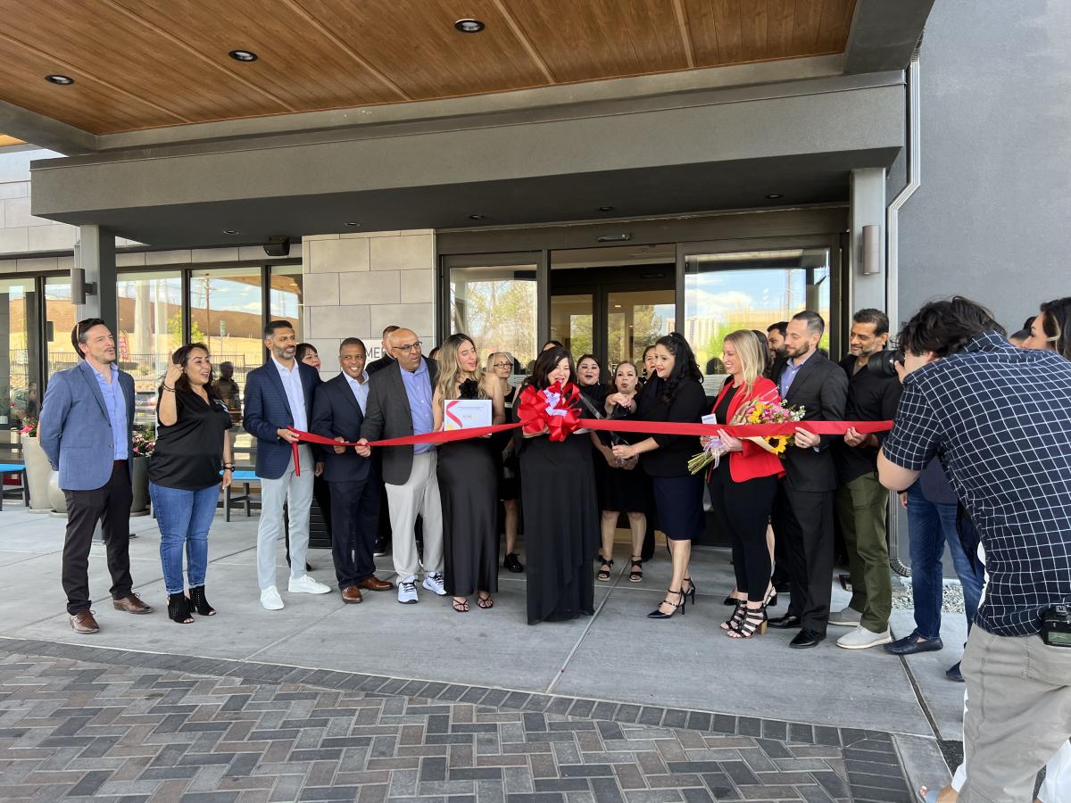 Many people stand in front of the Home2Suites Airport hotel behind a red ribbon to celebrate its grand opening.